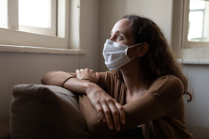 Young woman wearing a mask sitting indoors looking out the window, reflecting on bad things people face in life.