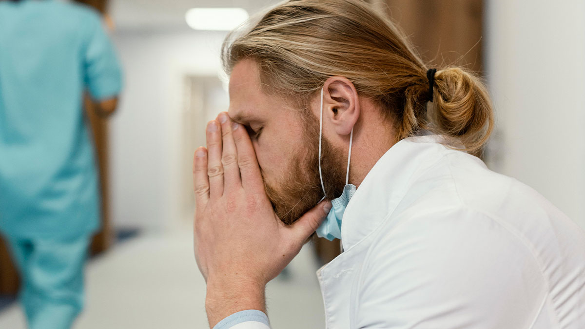 Stressed man in medical coat with mask lowered, sitting in hospital corridor, reflecting on slow and silent death risks.