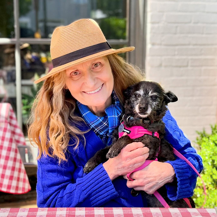 Beloved ’90s star and Rain Man co-star smiling outdoors in a blue sweater and hat holding a small black dog. Beloved ’90s star and Rain Man co-star smiling outdoors in a blue sweater and hat holding a small black dog.