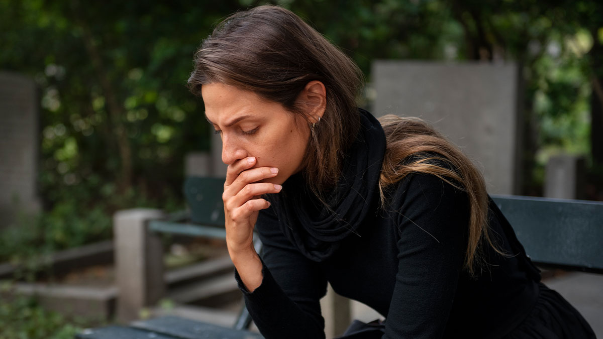 Woman in black sitting on bench outdoors looking distressed, reflecting drama involving best friend of late man and memorial plans