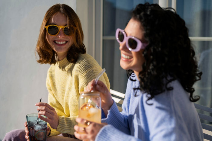 Two women wearing sunglasses, smiling and drinking beverages outdoors, reflecting friendship on the rocks and emotional distance.