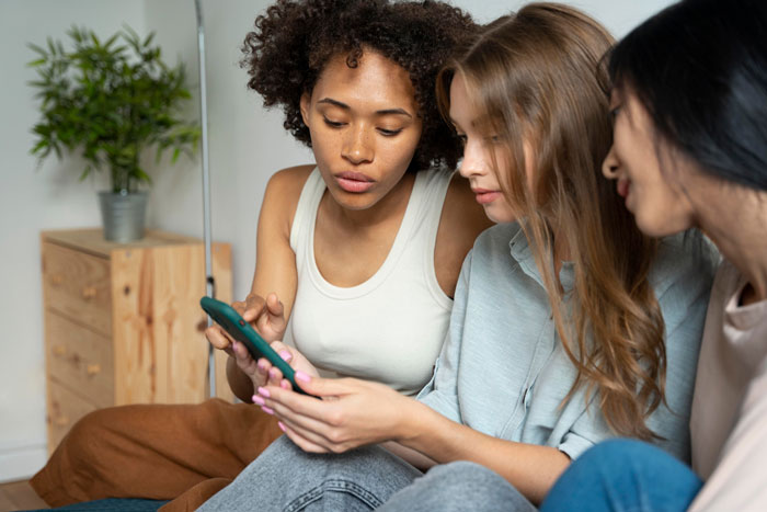 Three women sitting together looking at a phone, reflecting friendship of 10 years on the rocks and tension among friends. Three women sitting together looking at a phone, reflecting friendship of 10 years on the rocks and tension among friends.