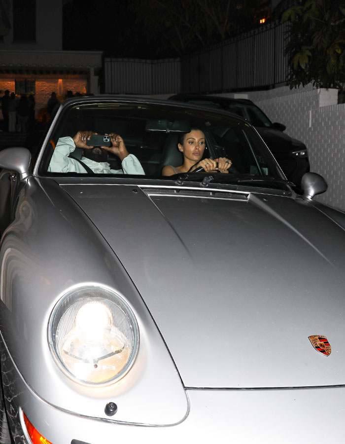 Bianca Censori driving a grey Porsche at night with a passenger using a smartphone inside the car. Bianca Censori driving a grey Porsche at night with a passenger using a smartphone inside the car.
