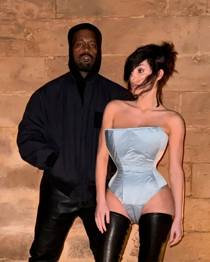 Kanye West standing next to a woman in a corset and thigh-high boots against a stone wall at night. Kanye West standing next to a woman in a corset and thigh-high boots against a stone wall at night.