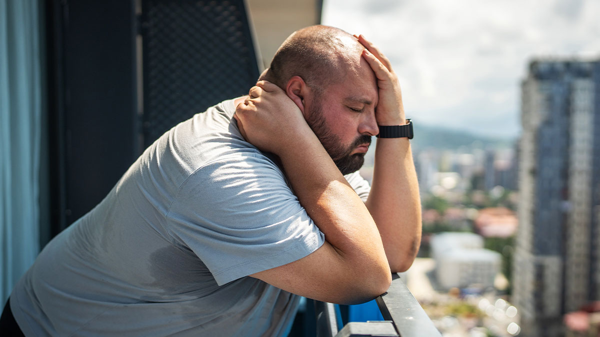 Man showing bil struggles on China trip, leaning on balcony railing with hand on forehead, looking stressed and tired.