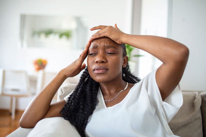 Teacher sitting on a couch with a pained expression, holding her head, stressed from paying family’s bills for years. Teacher sitting on a couch with a pained expression, holding her head, stressed from paying family’s bills for years.