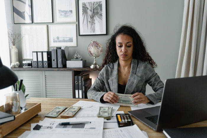 Woman teacher counting money at desk with bills and laptop, reflecting on paying family’s bills and feeling criticized. Woman teacher counting money at desk with bills and laptop, reflecting on paying family’s bills and feeling criticized.