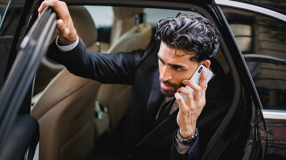 Man in black suit sitting in car talking on phone, distracted and ignoring lawyer girlfriendu2019s ceremony nearby.