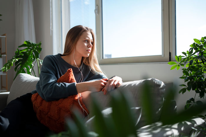 Young woman sitting on couch looking shocked and contemplative, reflecting on being called a downgrade by boyfriend’s mom.