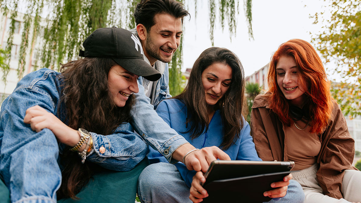 Group of friends breaking guy or girl code, smiling and interacting together while looking at a tablet outdoors.