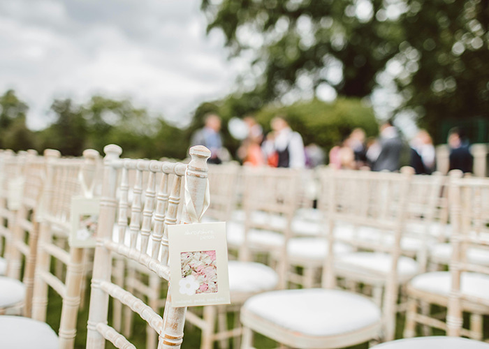 Rows of empty white chairs set up outdoors for a wedding ceremony with blurred guests in the background Rows of empty white chairs set up outdoors for a wedding ceremony with blurred guests in the background