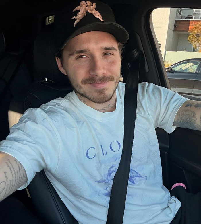 Brooklyn Beckham wearing a black cap and white shirt, sitting in a car with a calm expression amid family feud. Brooklyn Beckham wearing a black cap and white shirt, sitting in a car with a calm expression amid family feud.