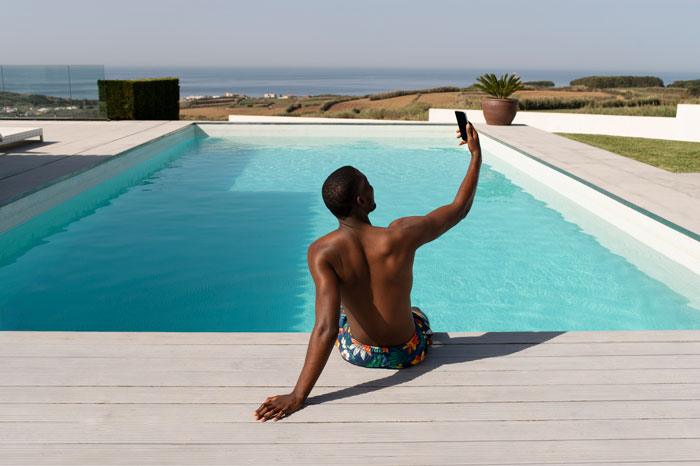Man sitting by pool taking selfie, illustrating man calling cops after bro and family broke into his home to use pool. Man sitting by pool taking selfie, illustrating man calling cops after bro and family broke into his home to use pool.