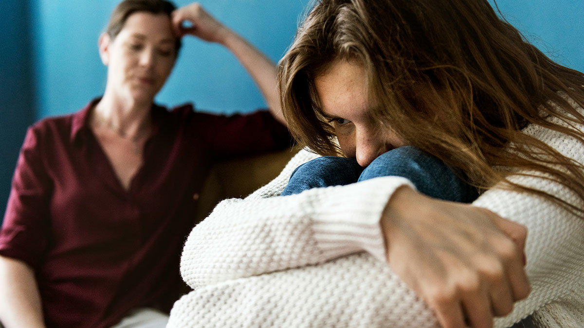 Daughter looking upset hugging knees while mother sits behind her frustrated, reflecting family relationship conflict.