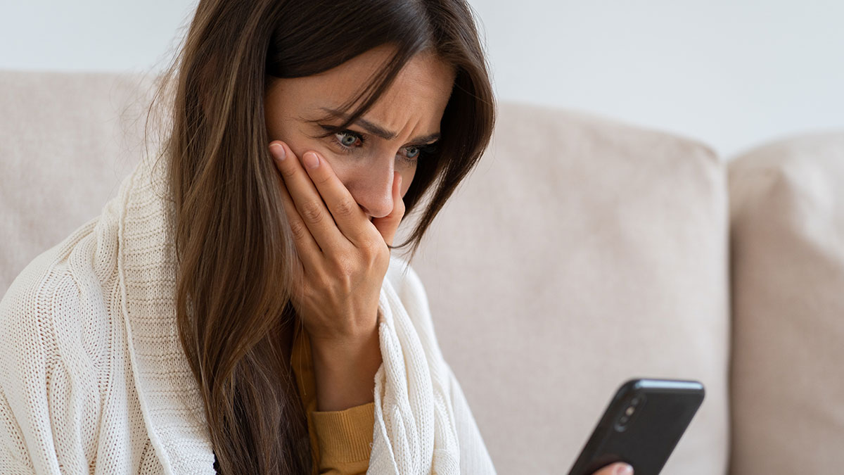 Woman looking shocked at phone screen on couch, reacting to man hiding a secret second family revealed by phone.