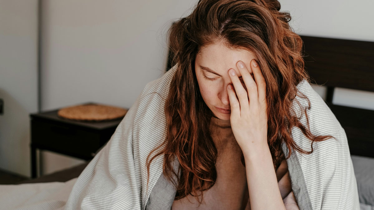 Worried childfree woman with hand on face sitting on bed wrapped in blanket, skipping trip to avoid babysitting on vacation