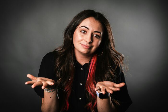 Young woman with dark hair and red streaks shrugging, expressing subtle signs of low self-esteem in a neutral background studio.