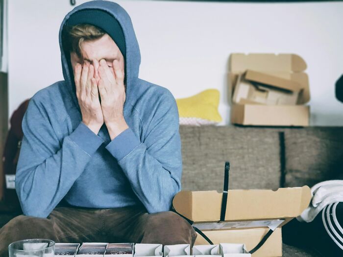 Young man in a hoodie sitting on a couch covering his face, showing subtle signs of low self-esteem and distress.