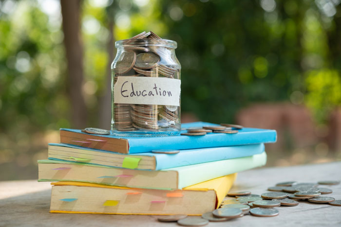 Glass jar labeled education filled with coins on colorful stacked books outdoors representing saving money. Glass jar labeled education filled with coins on colorful stacked books outdoors representing saving money.