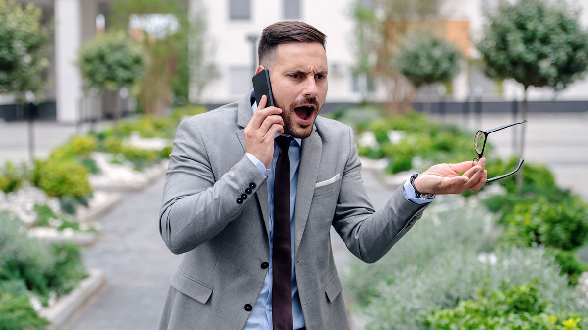 Man in gray suit speaking loudly on phone outdoors, holding glasses and looking frustrated in a public space.