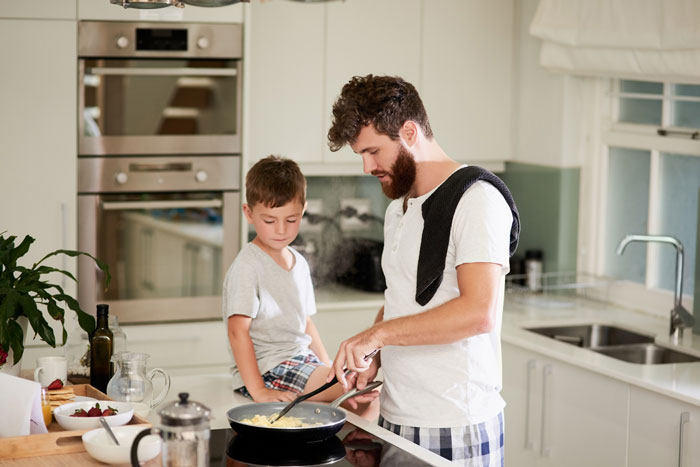 Man cooking breakfast for kids in modern kitchen while young boy watches, highlighting playing chef for her kids concept. Man cooking breakfast for kids in modern kitchen while young boy watches, highlighting playing chef for her kids concept.