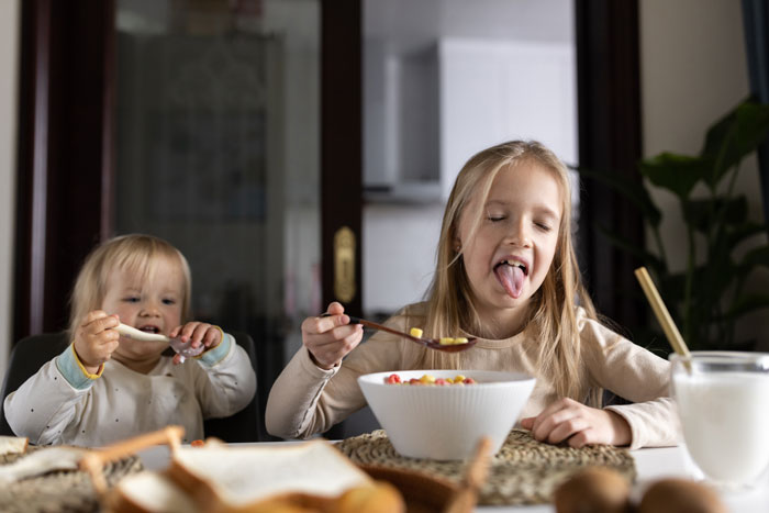 Two children eating breakfast cereal at a table while their uncle plays chef for them in a home kitchen. Two children eating breakfast cereal at a table while their uncle plays chef for them in a home kitchen.