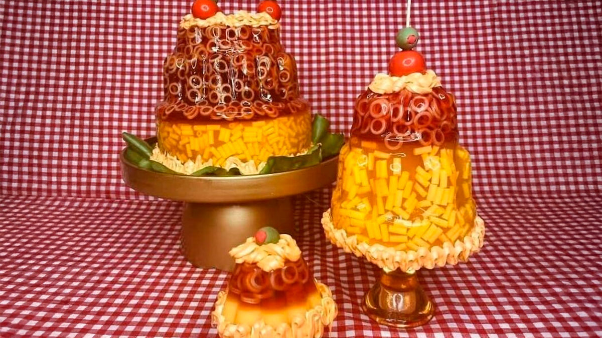 Layered jello mold with macaroni, cheese, and olives on cake stands against a red checkered background, gross foods concept.