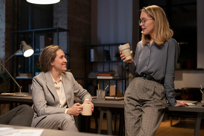 Two women in a corporate office having a casual conversation over coffee, illustrating ways to call out corporate pals' lies.