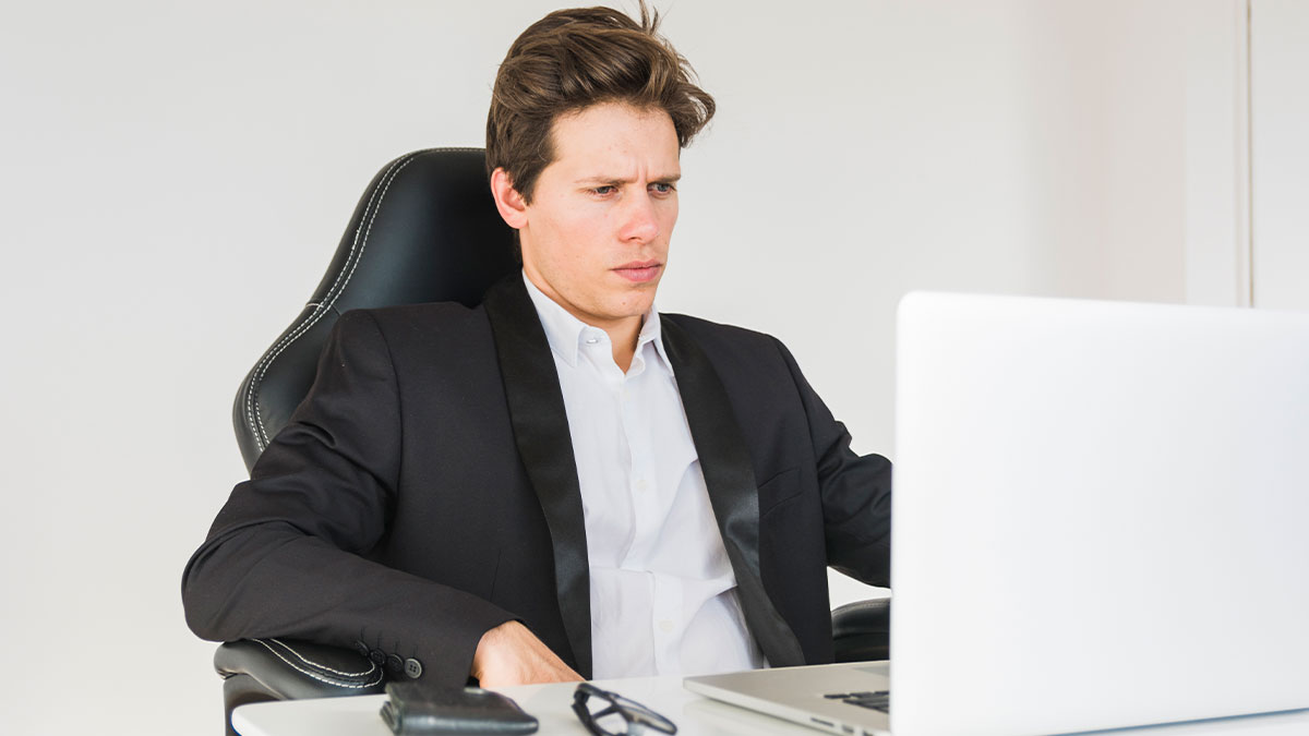 Young man in a black suit sitting at desk looking skeptical while using laptop, calling out corporate palsu2019 lies.