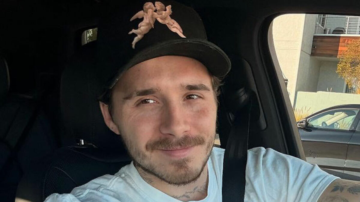 Brooklyn Beckham wearing a black cap with cherub design, sitting inside a car, looking slightly to the side and smiling.