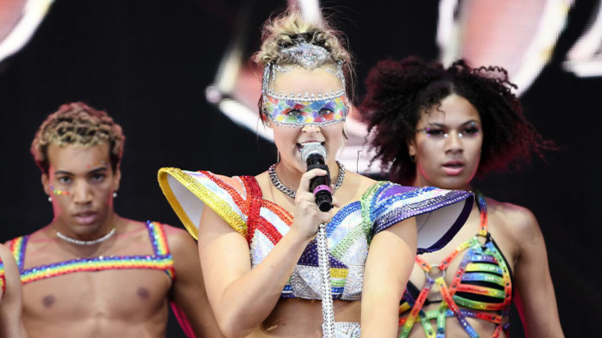 JoJo Siwa performing on stage with dancers, wearing colorful futuristic costumes and a decorated face mask.