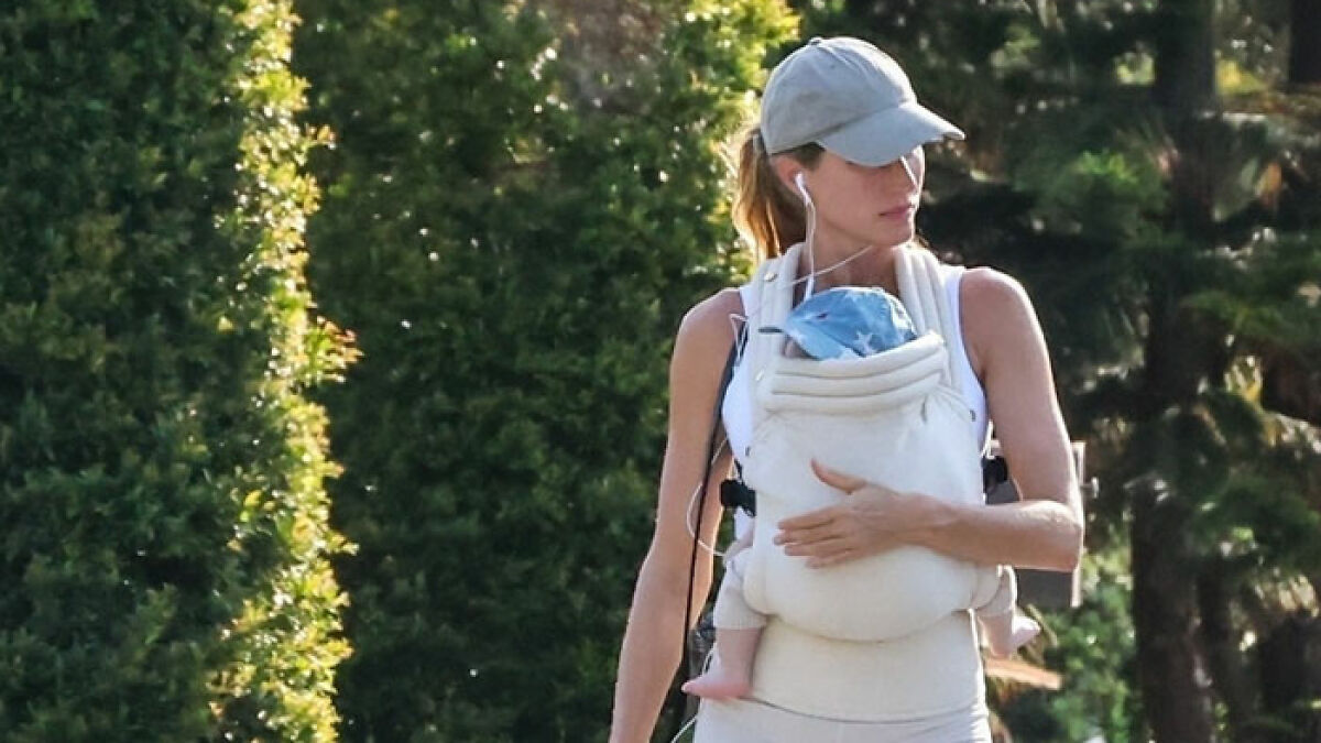 Gisele Bundchen walking baby in a carrier while walking the dog, wearing a cap and earbuds outdoors in sunlight.