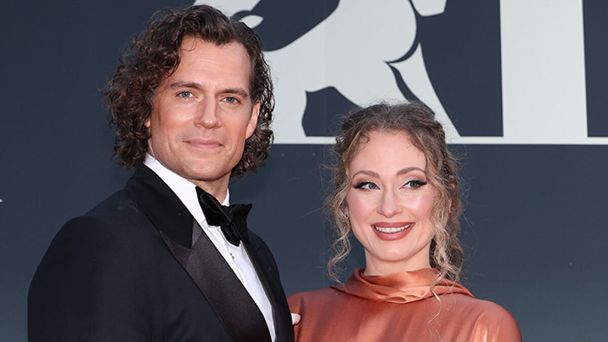 Henry Cavill with curly highlander hairstyle posing in black tuxedo alongside a woman in a satin copper dress at event.