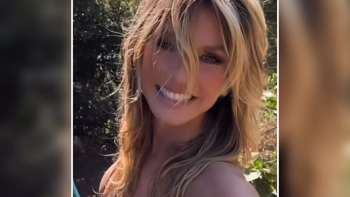 Heidi Klum smiling in a tiny bikini outdoors, filming a racy video in her garden on a sunny day.