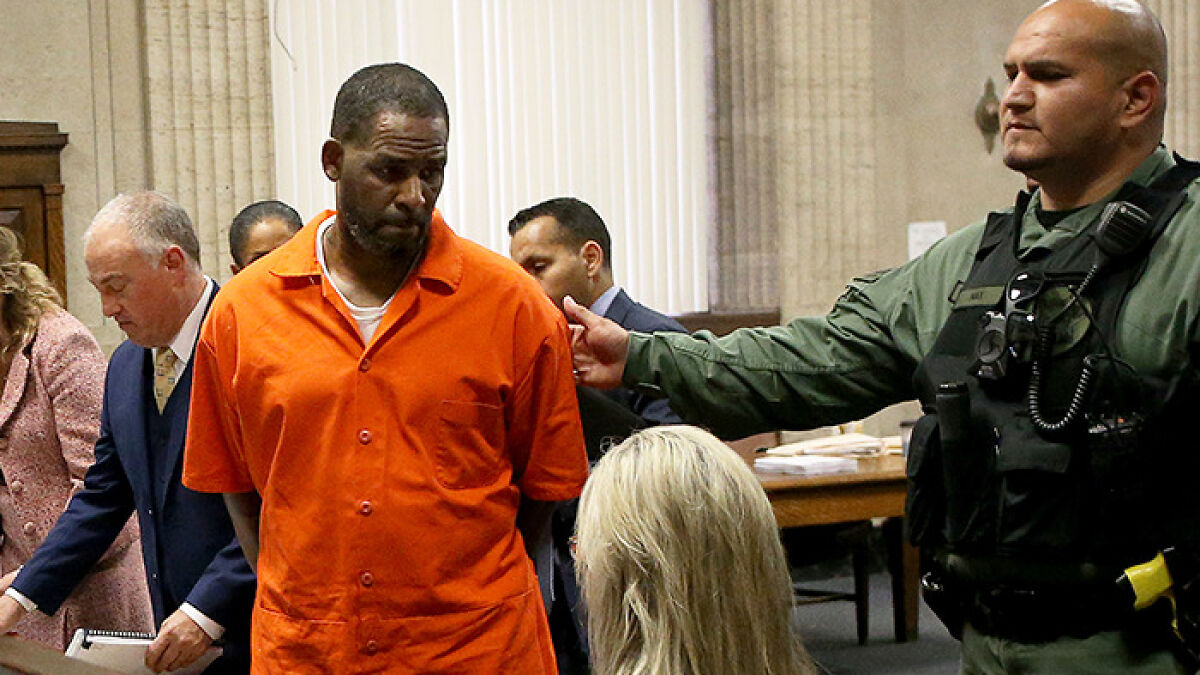 Rapper R Kelly in orange prison attire escorted by guard in a courtroom setting during legal proceedings.