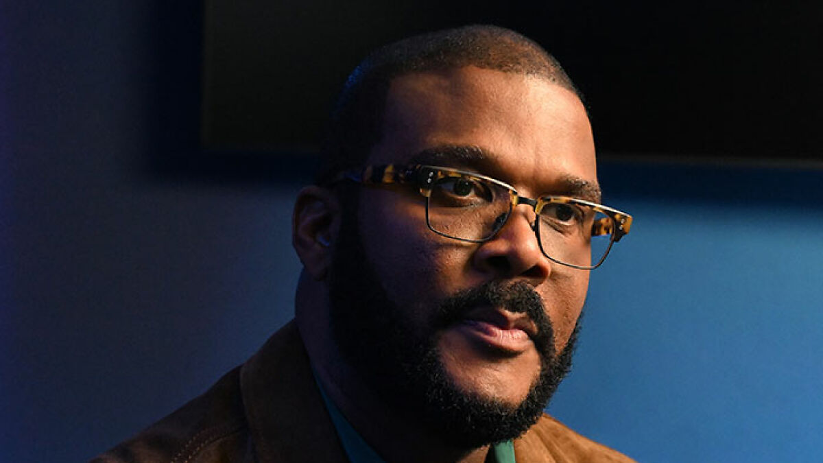 Tyler Perry wearing glasses and a brown jacket, looking serious against a dark blue background in an indoor setting