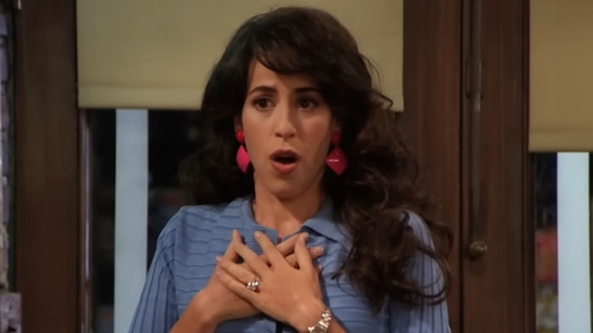 Actress from Friends with dark hair, wearing blue top and pink earrings, expressing surprise in a sitcom scene.