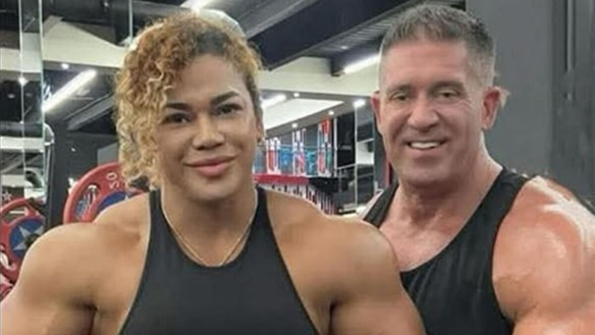 Woman bodybuilder with curly hair and man, both muscular and smiling, posing in a gym setting.
