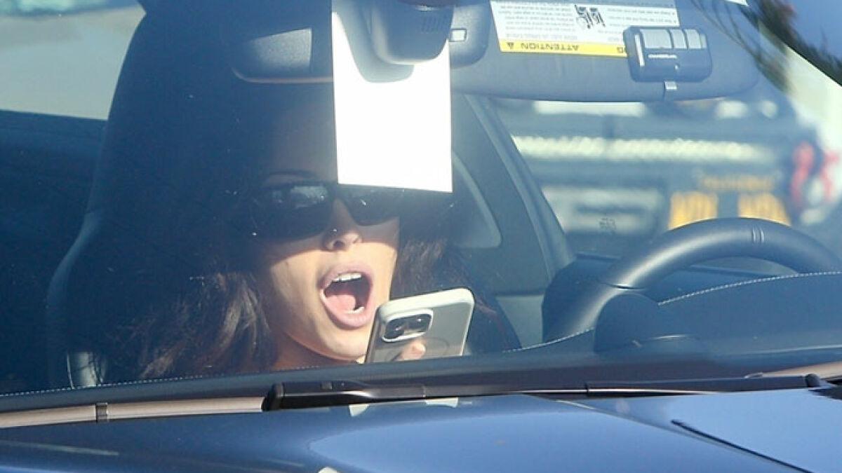 Bianca Censori in Porsche wearing sunglasses, holding phone, caught on camera for alleged law infringement while driving.
