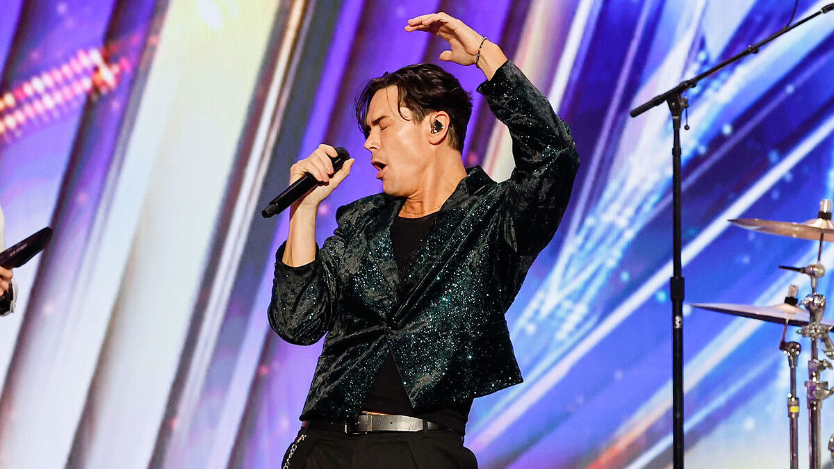 Tom Sandoval performing on Americas Got Talent stage, singing passionately with a glittery jacket and microphone.