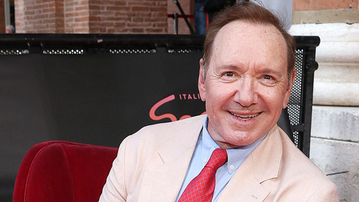 Kevin Spacey at an event in a beige suit and red tie, fueling plastic surgery rumors after his red carpet appearance.