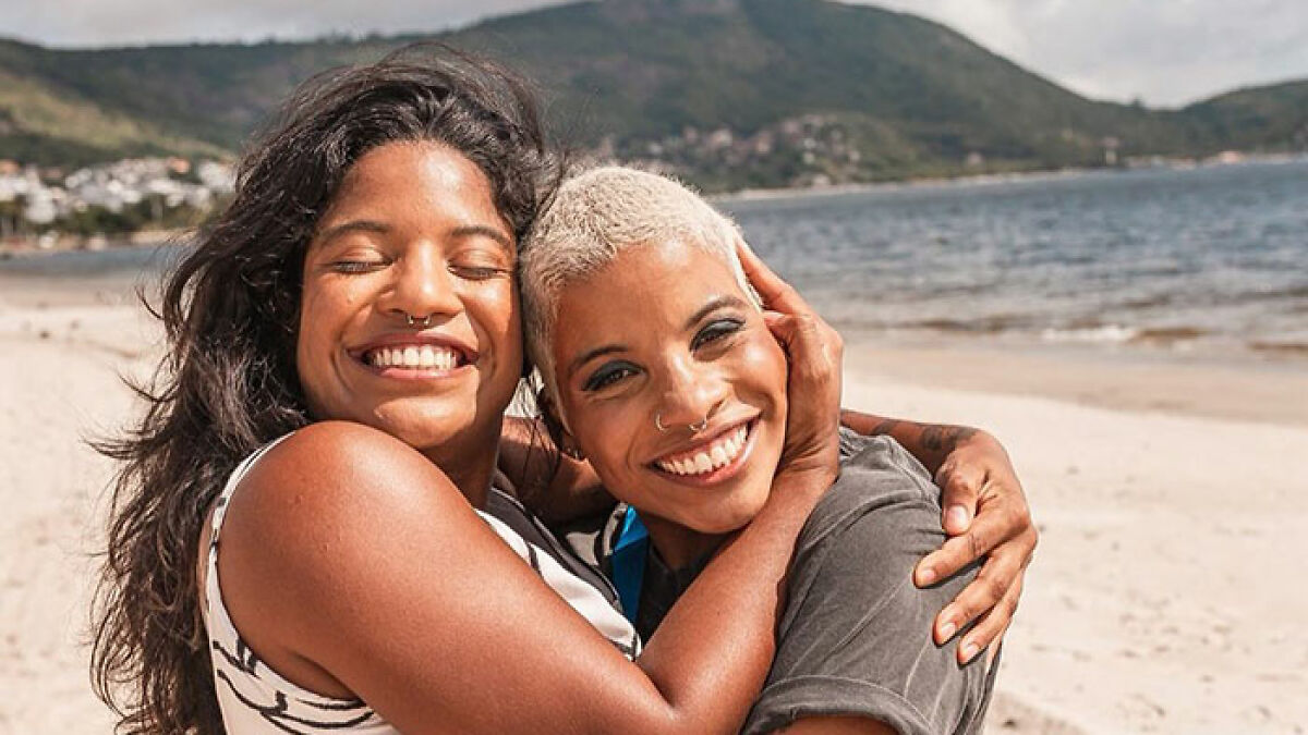 Two women smiling and hugging on a beach with a mountain and ocean in the background, related to volcano guide topic.