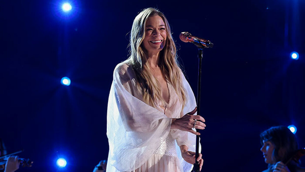 LeAnn Rimes performing on stage, smiling and holding a microphone, with blue stage lights in the background.