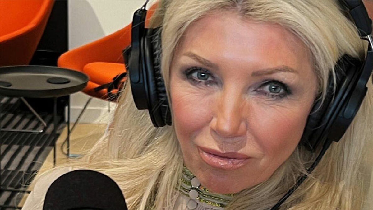 Blonde woman wearing headphones speaking into a microphone in a room with orange chairs, focused on anti-vax mom topic.