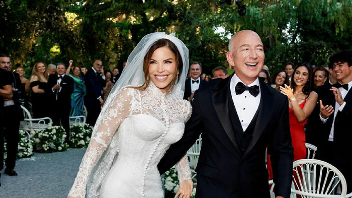 Lauren Su00e1nchez in a white lace wedding dress with blue veins on her hands, smiling beside a man in a black tuxedo outdoors.