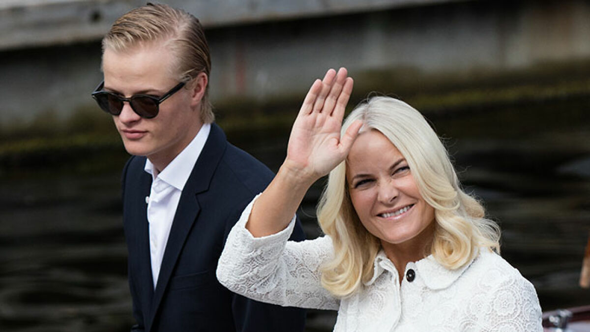 Norwegian Crown Princessu2019s son wearing sunglasses beside her, with the princess waving and smiling outdoors by the water.