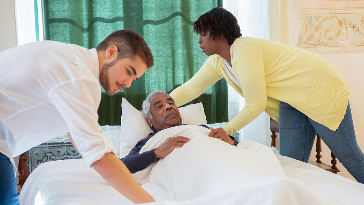 Two coworkers adjusting an elderly man in bed, discussing how to report diabolical things to HR at work.