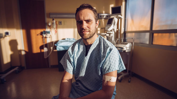 Man in hospital gown sitting in a medical room with bandage on arm, relating to colleague sick and stalking story. Man in hospital gown sitting in a medical room with bandage on arm, relating to colleague sick and stalking story.