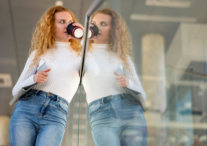Woman with red hair drinking coffee and holding tablet, reflecting on glass wall, symbolizing stalking colleague to confirm sickness. Woman with red hair drinking coffee and holding tablet, reflecting on glass wall, symbolizing stalking colleague to confirm sickness.