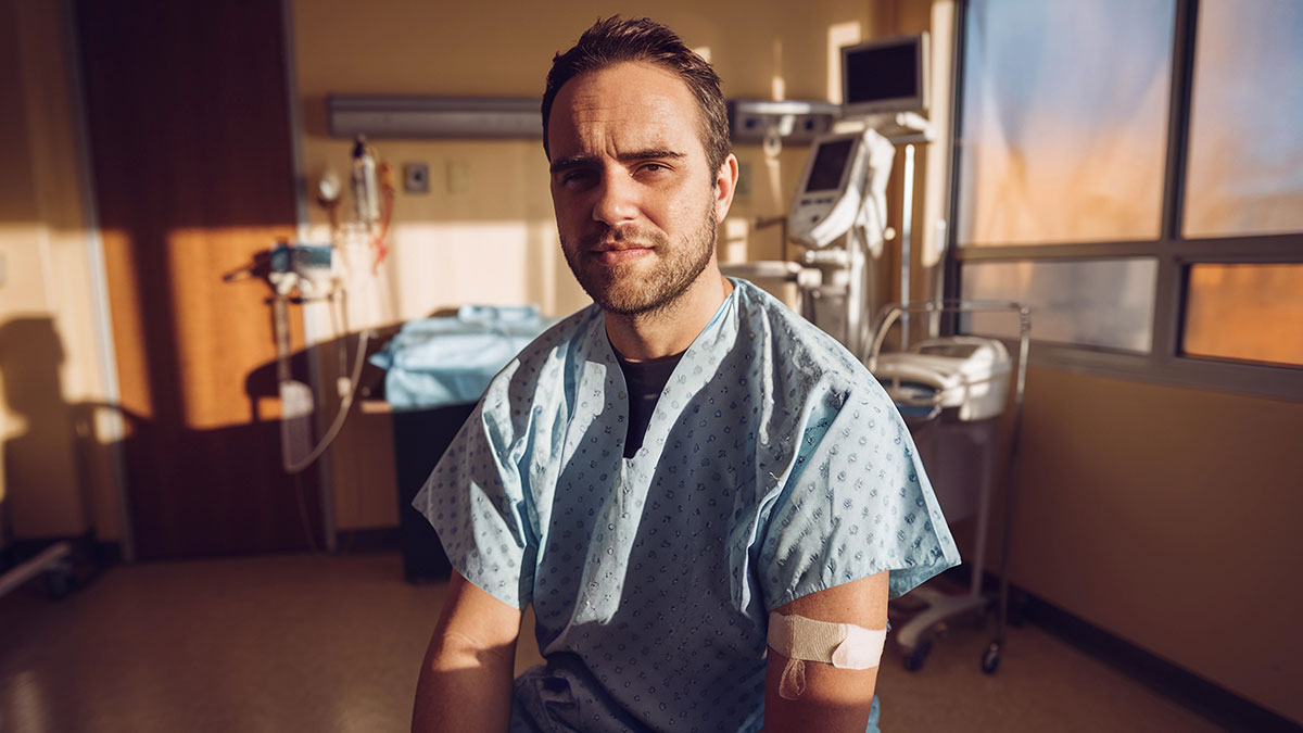Man in hospital gown with bandaged arm sitting in medical room, depicting colleague confirmed sick in hospital setting